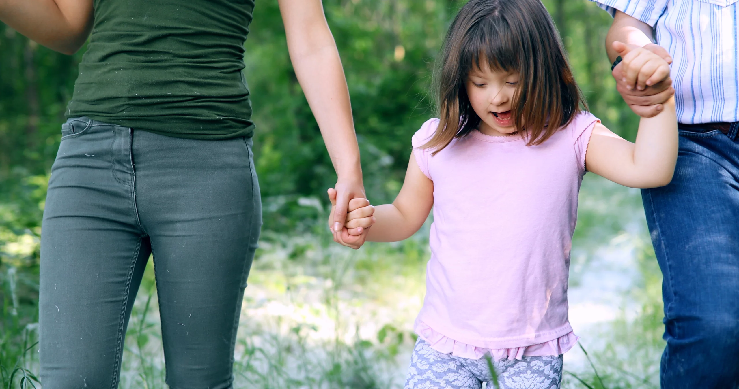 A young girl in a pink shirt holds hands with two adults while walking through a sunlit forest, symbolizing care and financial security
